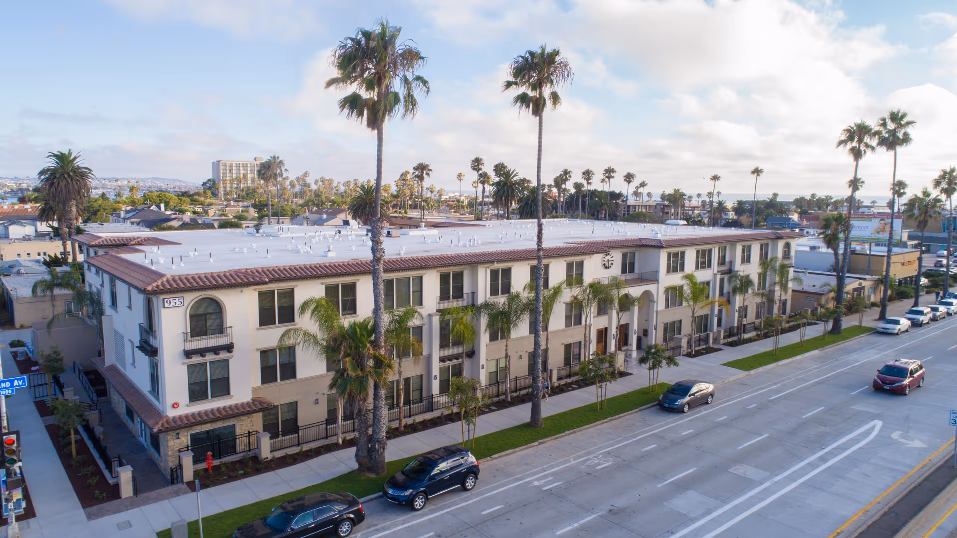 Aerial view of a two-story senior living facility building with a beige exterior and red-tiled roof, lined with palm trees along the sidewalk. Several cars are parked on the street in front of the building, and the surrounding area includes other buildings and more palm trees under a partly cloudy sky.