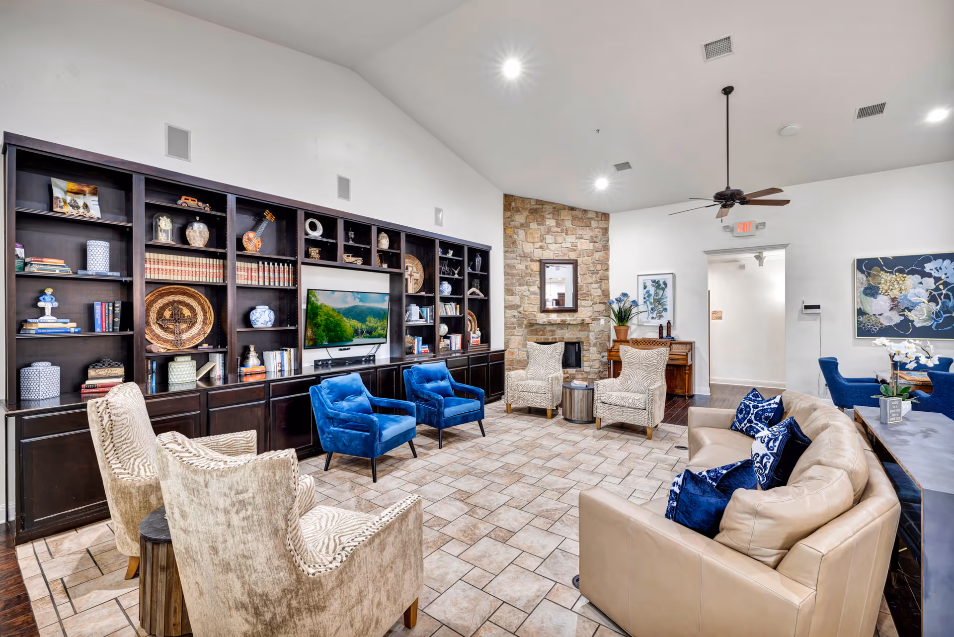 Spacious lounge area with beige seating, blue chairs, built-in dark bookshelf, TV, fireplace, and accent wall.