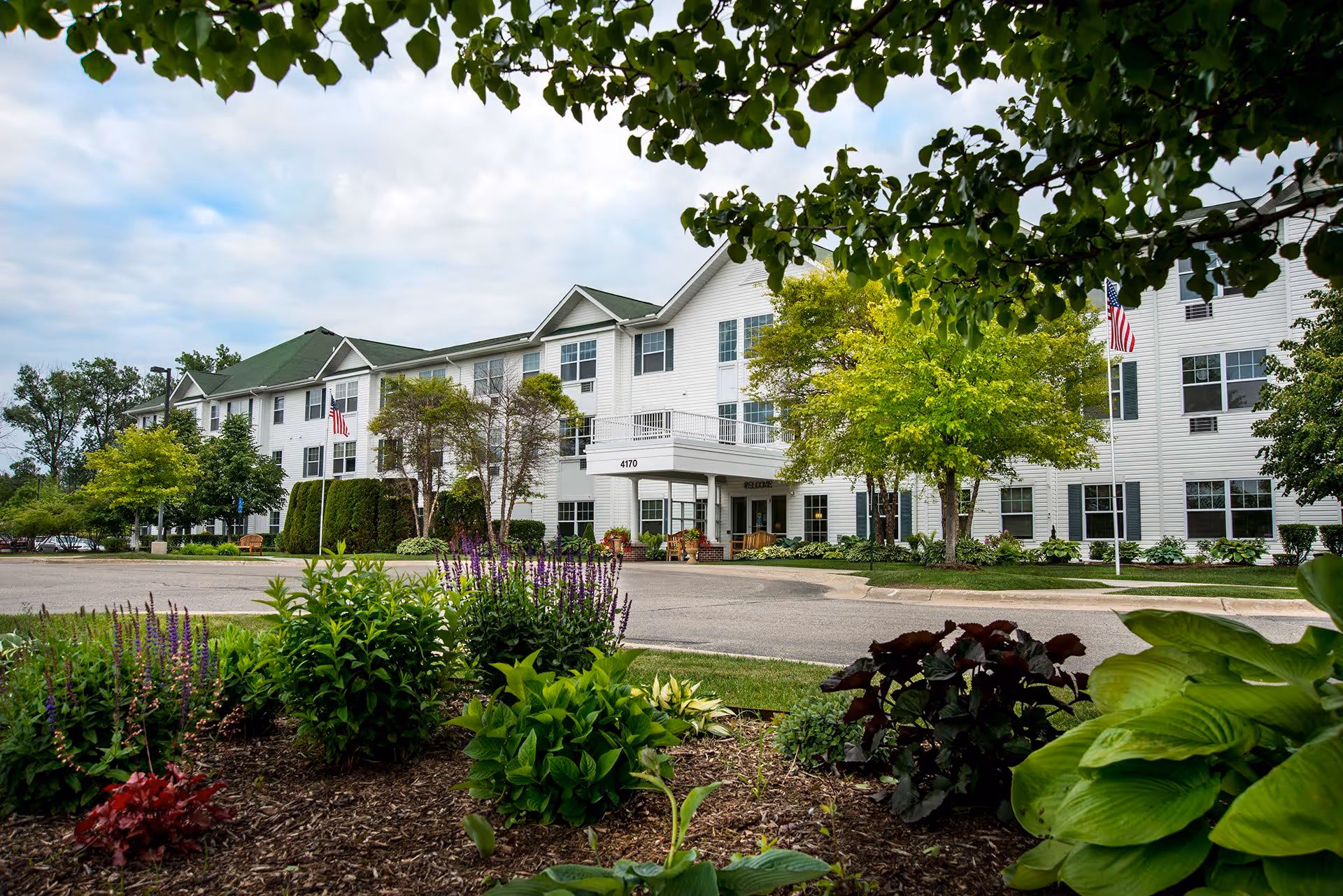 Exterior view of Mercy Village, a three-story white building with green roofs, surrounded by trees and landscaped garden beds with various plants and flowers. An American flag is visible near the entrance, and the building number 4170 is displayed above the main entrance.