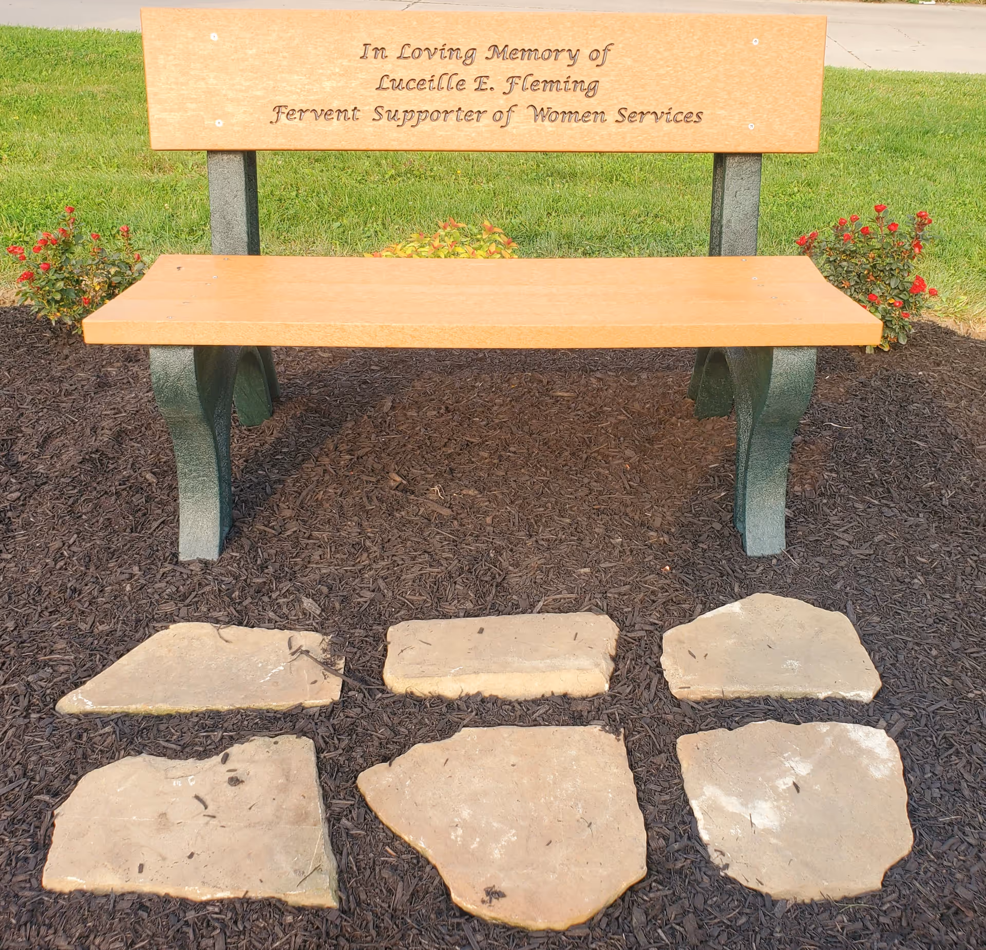 Outdoor memorial bench with engraved plaque on mulch beside stepping-stone path
