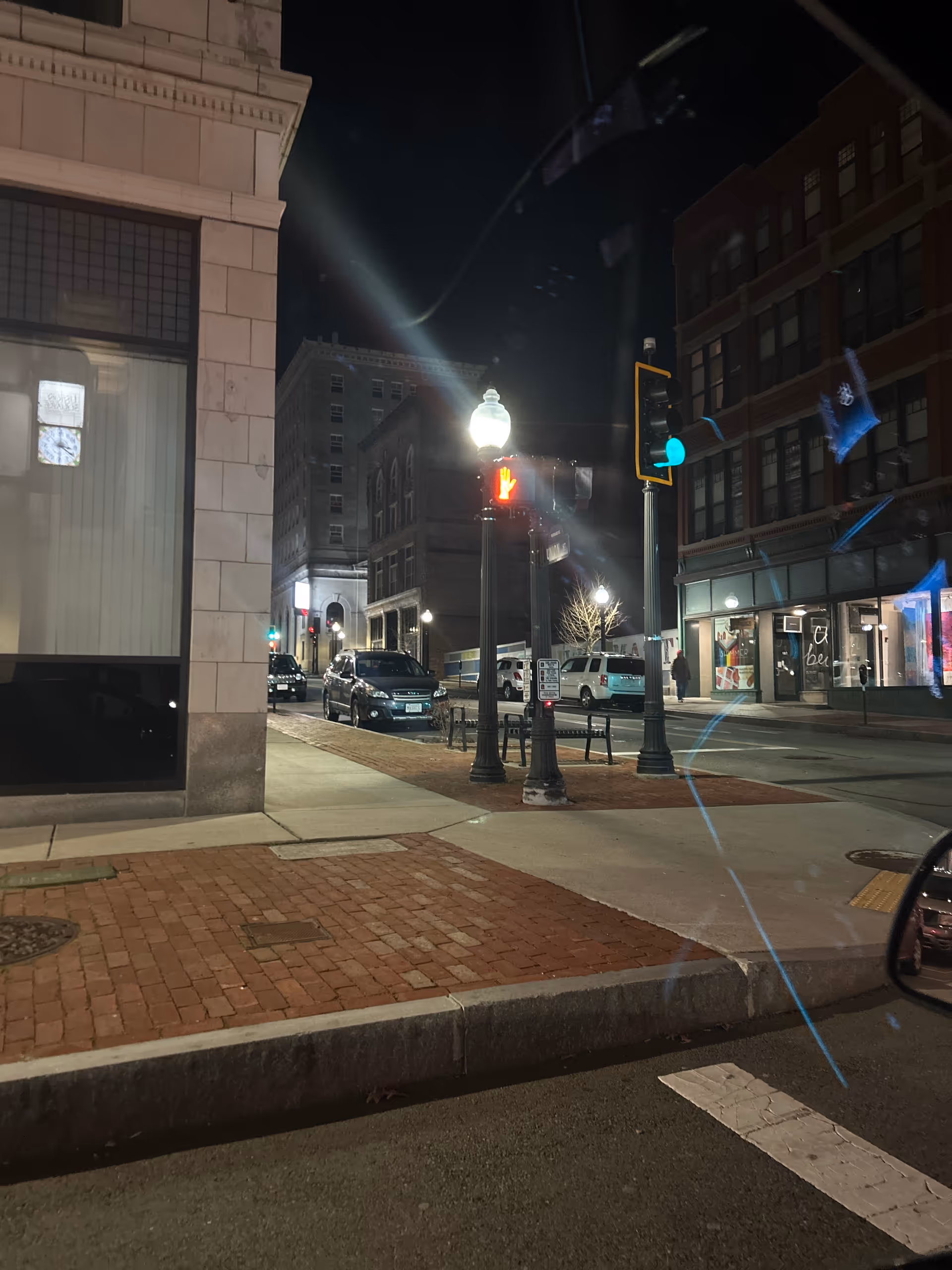 Night city street scene with brick sidewalk, lampposts, and storefronts along the curb.