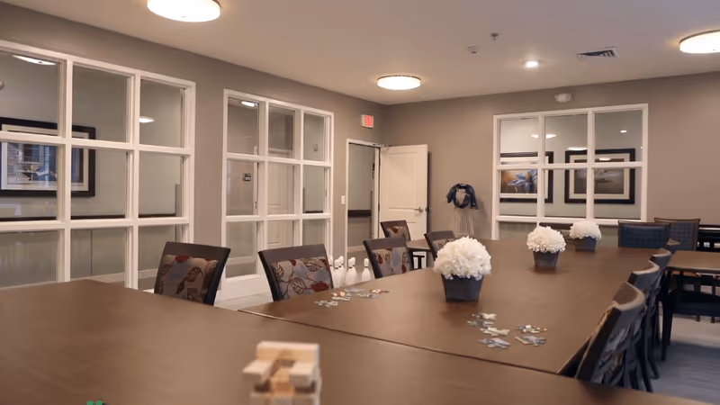A spacious, well-lit room with a large wooden table surrounded by chairs. The table has small flower arrangements and puzzle pieces scattered on it. The walls are beige with large windows and framed artwork. There is a door at the back and a coat rack with a jacket hanging on it.