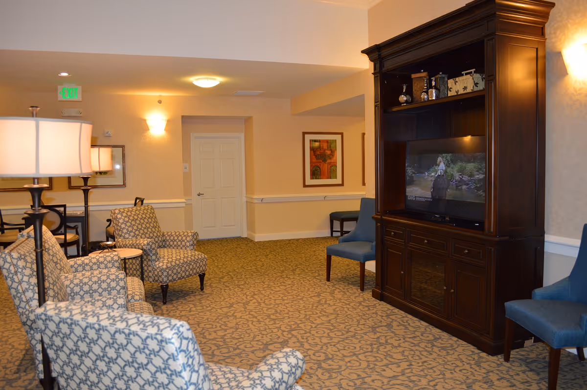 A cozy living room area in a senior living facility with patterned armchairs, blue chairs, a dark wooden entertainment center with a TV, soft lighting, and framed artwork on the walls.