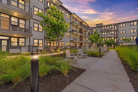 Exterior courtyard of Sage Plum Creek senior living with walkway, seating, trees, and a multi-story building.