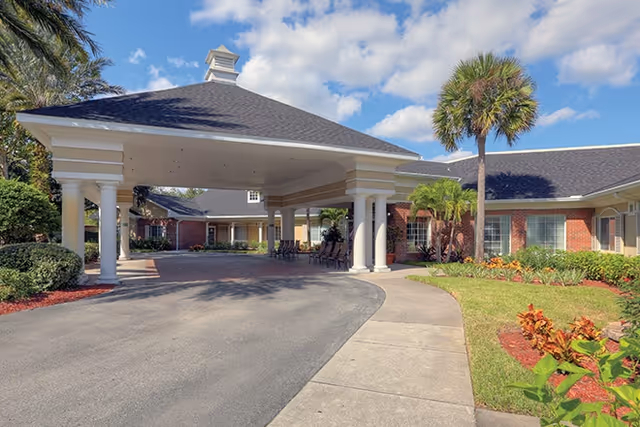 Covered front entrance porte-cochère with white columns, brick building, palm trees, and landscaped driveway under a blue sky.