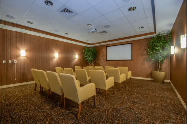 Small screening room with rows of beige upholstered chairs facing a blank projection screen and potted plants.