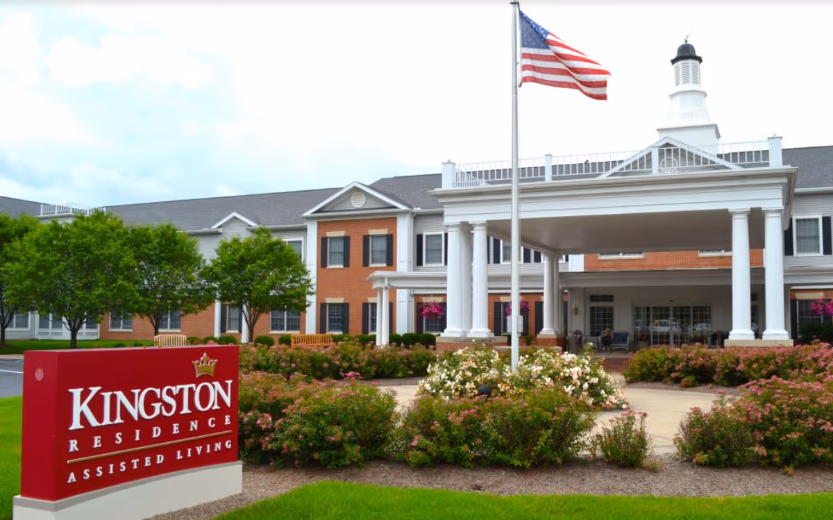 Front entrance of Kingston Residence assisted living with a covered portico, American flag, landscaped flowerbeds, and a red Kingston Residence sign.