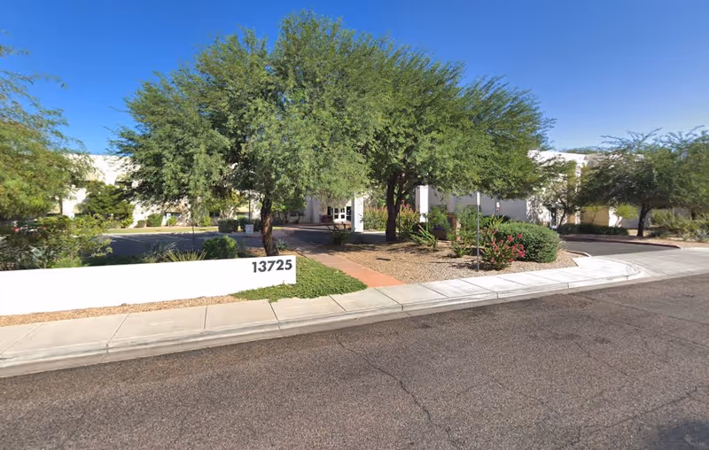 Front entrance of a single-story assisted living facility with trees, landscaping, a walkway, and a white address wall reading 13725.
