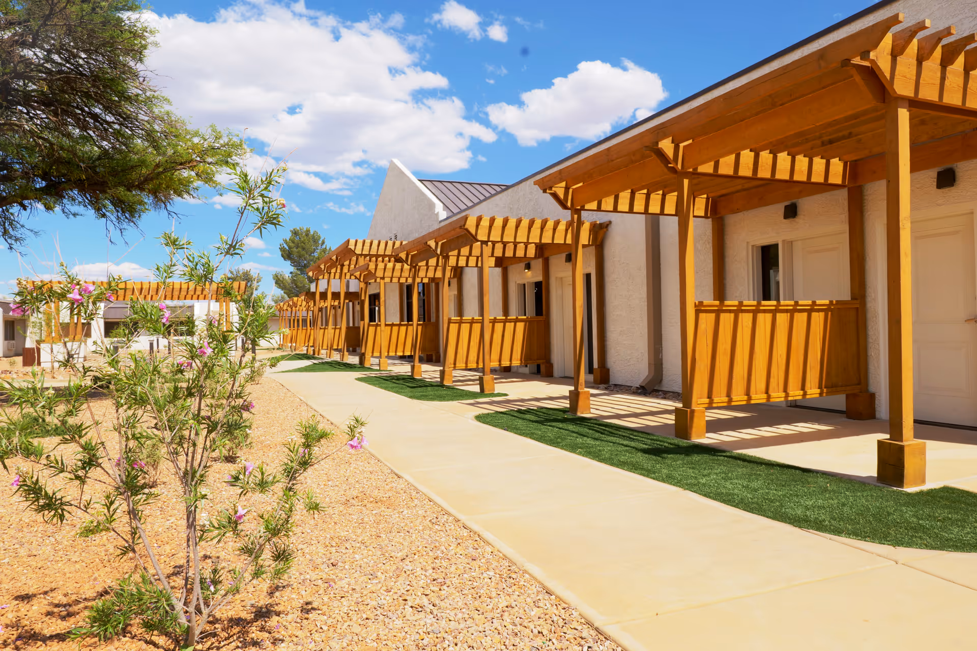 Row of single-story senior living units with wooden pergola-covered porches along a paved walkway and desert landscaping.