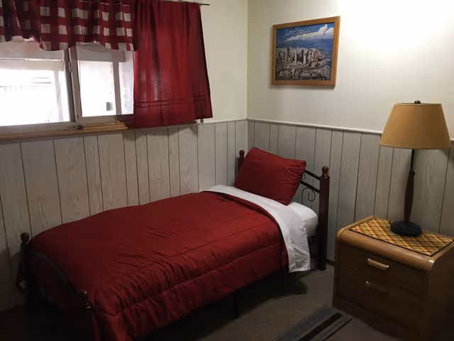 A small bedroom with a single bed dressed in red bedding, a wooden nightstand with a lamp, wood-paneled lower walls, a window with red curtains and a framed picture on the wall.