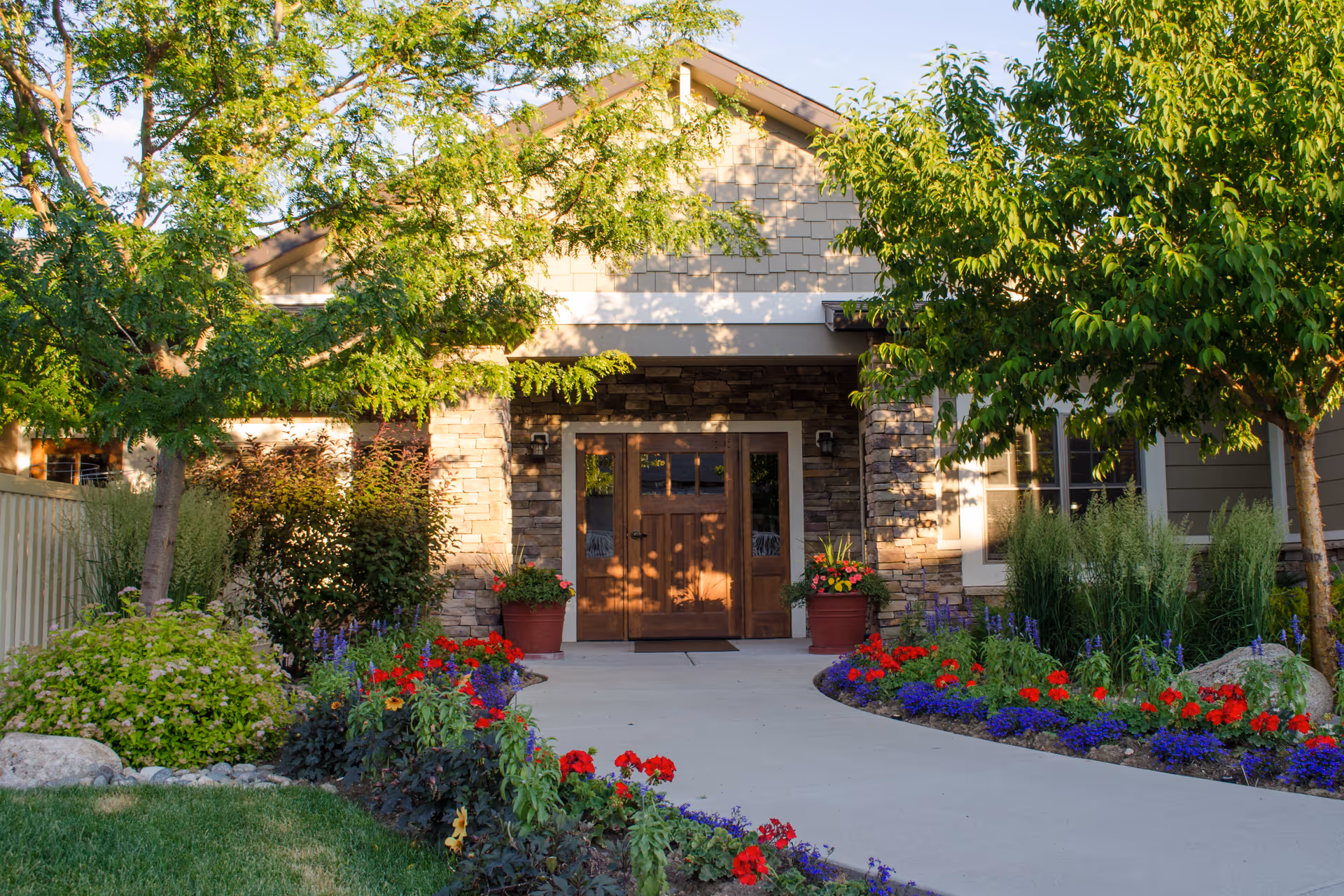 Front entrance of a building with a stone facade and wooden double doors, surrounded by a well-maintained garden with colorful flowers and green trees on either side of a curved concrete pathway.