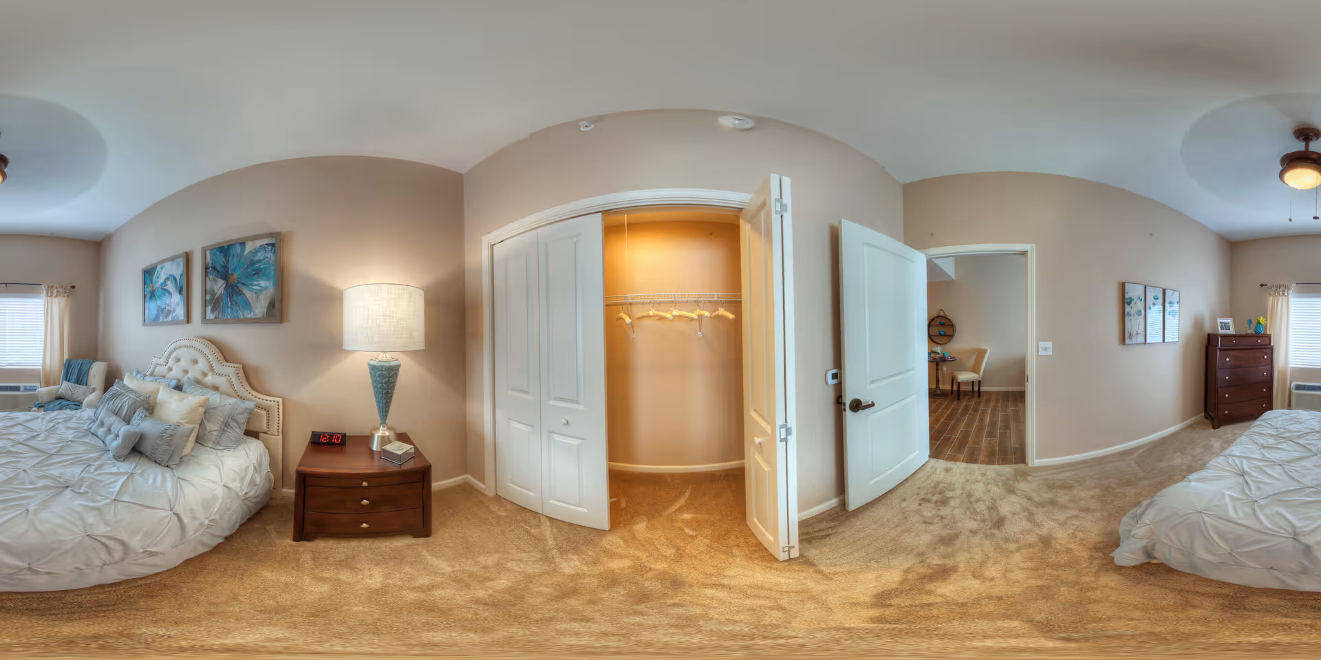 Panoramic view of a furnished bedroom with a bed, nightstand and lamp, open closet, and doorway to an adjoining room.