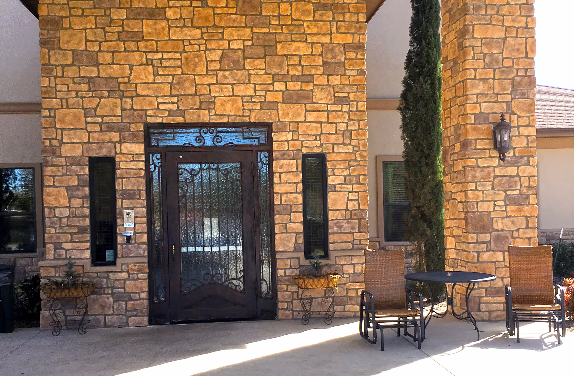 Stone exterior entrance of a building with a decorative wrought iron door, two narrow windows on either side, two wicker chairs, a small round table, and tall greenery near the entrance.