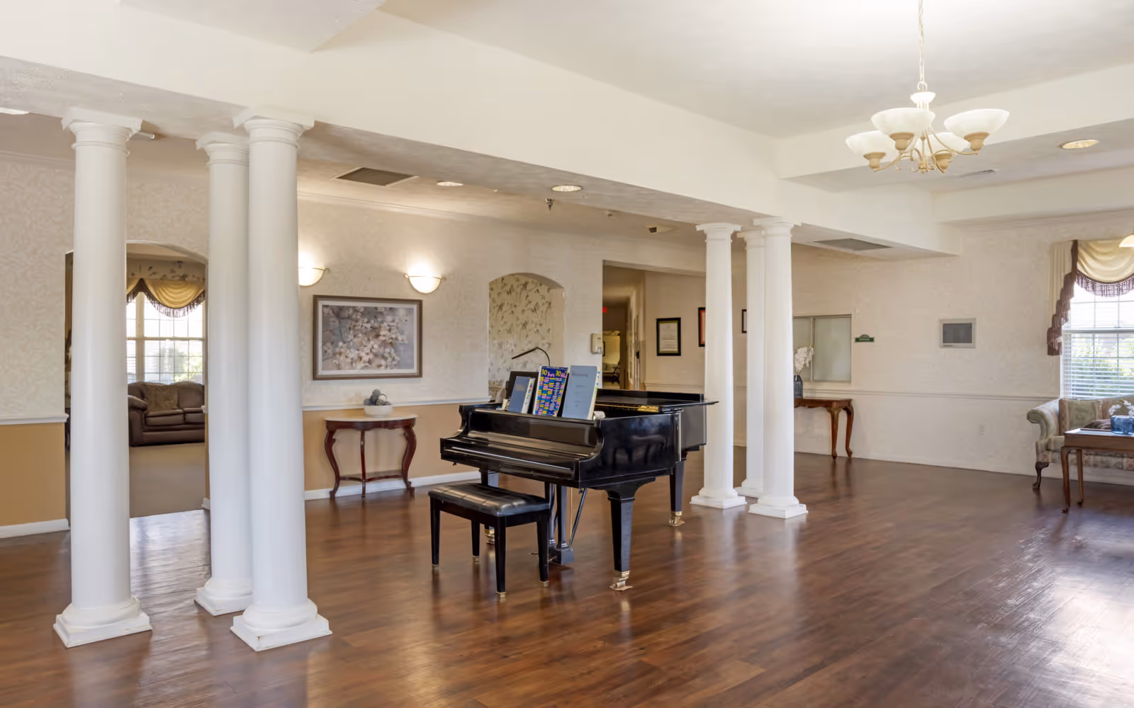 A spacious room with wooden flooring featuring a black grand piano and matching bench in the center. The room has white decorative columns, beige walls with floral wallpaper, and soft lighting from wall sconces and a chandelier. There is a small wooden table with a decorative bowl and framed artwork on the wall. In the background, there are windows with draped curtains and a seating area with a sofa and a wooden coffee table.