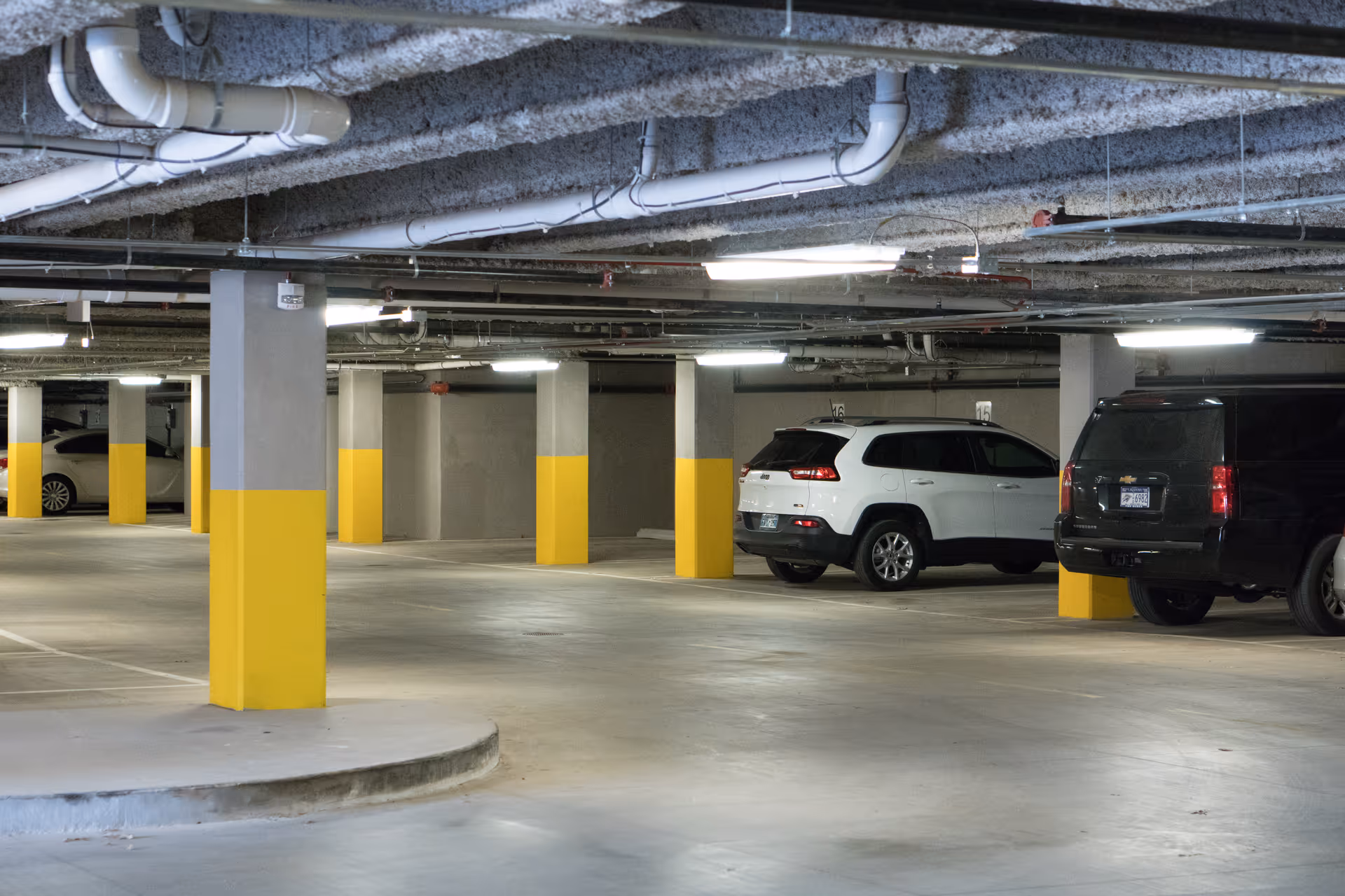 Indoor parking garage with concrete pillars painted gray and yellow, several parked cars including a white SUV and a black van, and overhead fluorescent lighting.