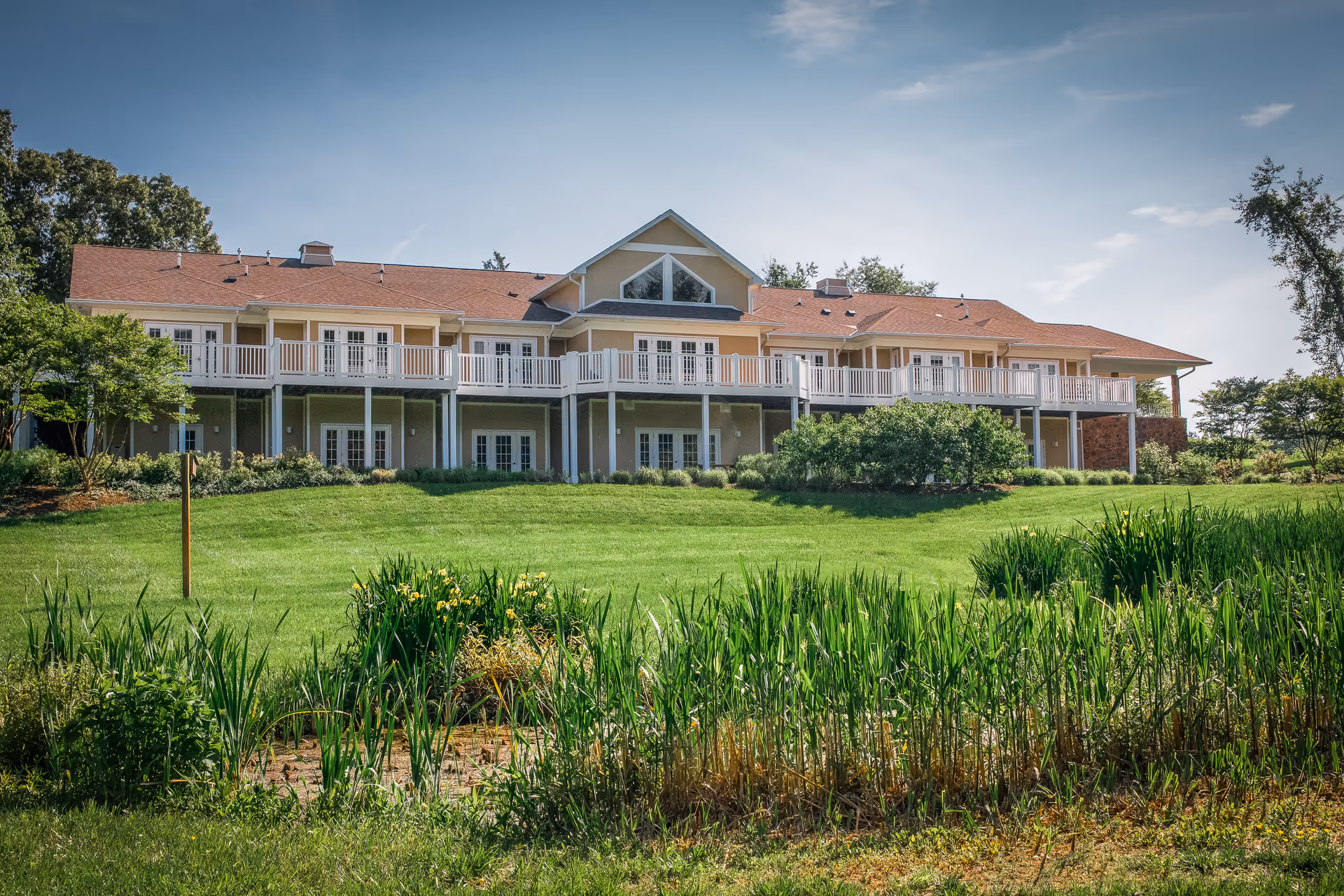 A large, two-story hospice house with a long white balcony, well-kept lawn, and pond in foreground.