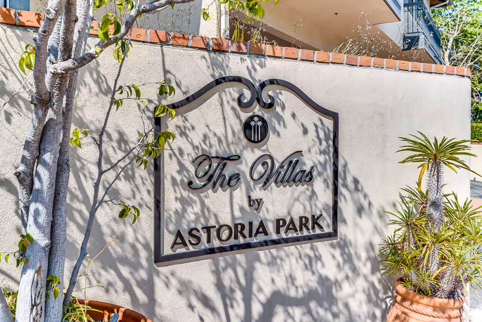 A decorative wall sign reading "The Villas by ASTORIA PARK" mounted on a stucco entrance wall with potted plants and tree shadows.
