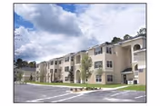 Beige multi-story senior living building with arched entry, parking lot, and blue sky.