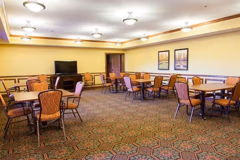 A spacious room with multiple tables and chairs arranged for group seating, featuring patterned carpet, beige walls with wooden trim, ceiling lights, a flat-screen TV on a stand, and two framed landscape paintings on the wall.