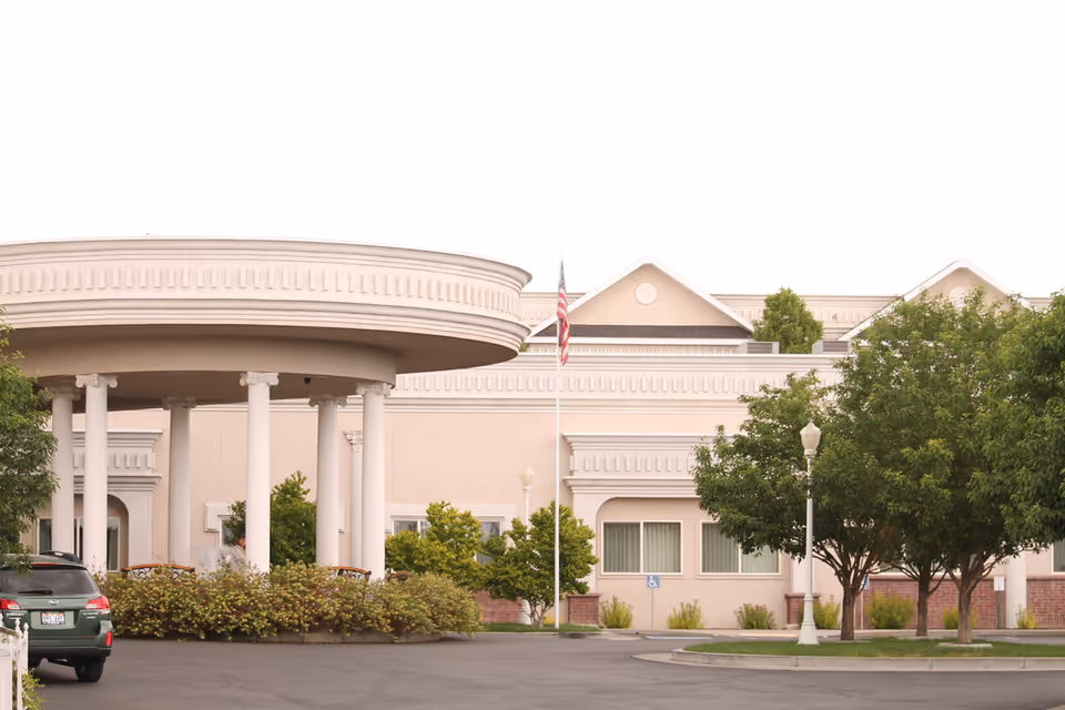 Front entrance of a senior living facility with a circular porte-cochere supported by columns, a flagpole, landscaping, and a parked car.