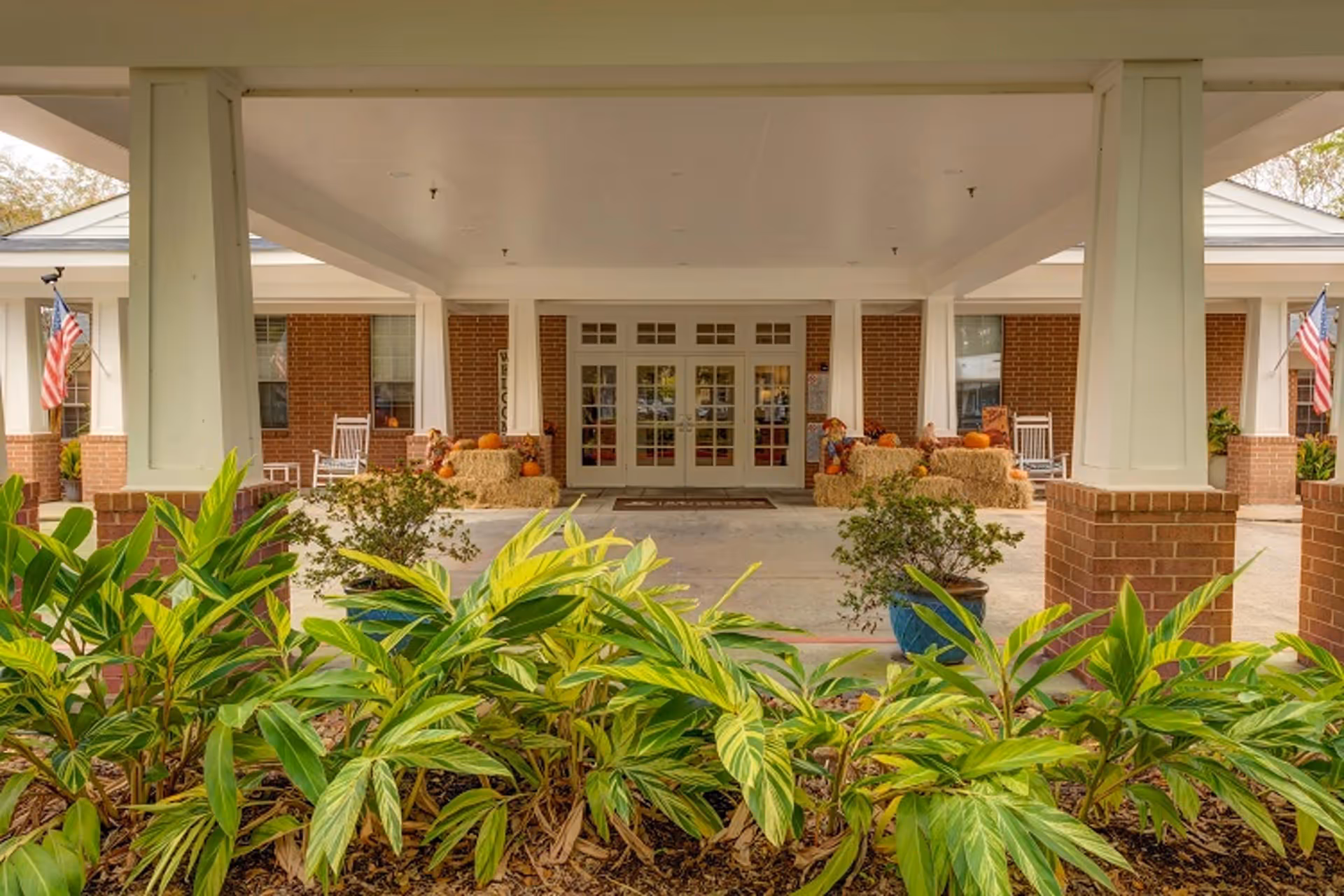 Covered front entrance of a senior living facility with columns, potted plants and landscaping in the foreground, rocking chairs, fall decorations and double glass doors under a portico.