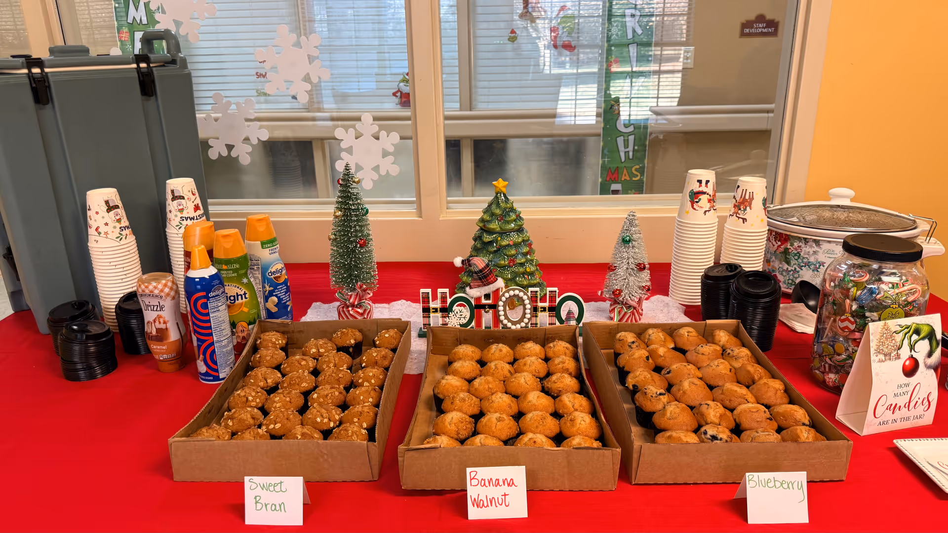 Holiday snack table with three boxes of muffins, drinks, cups, and festive décor.