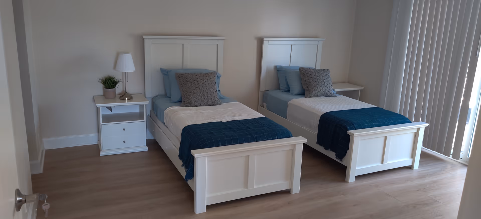 Two white twin beds with blue bedding and a nightstand in a bright bedroom.