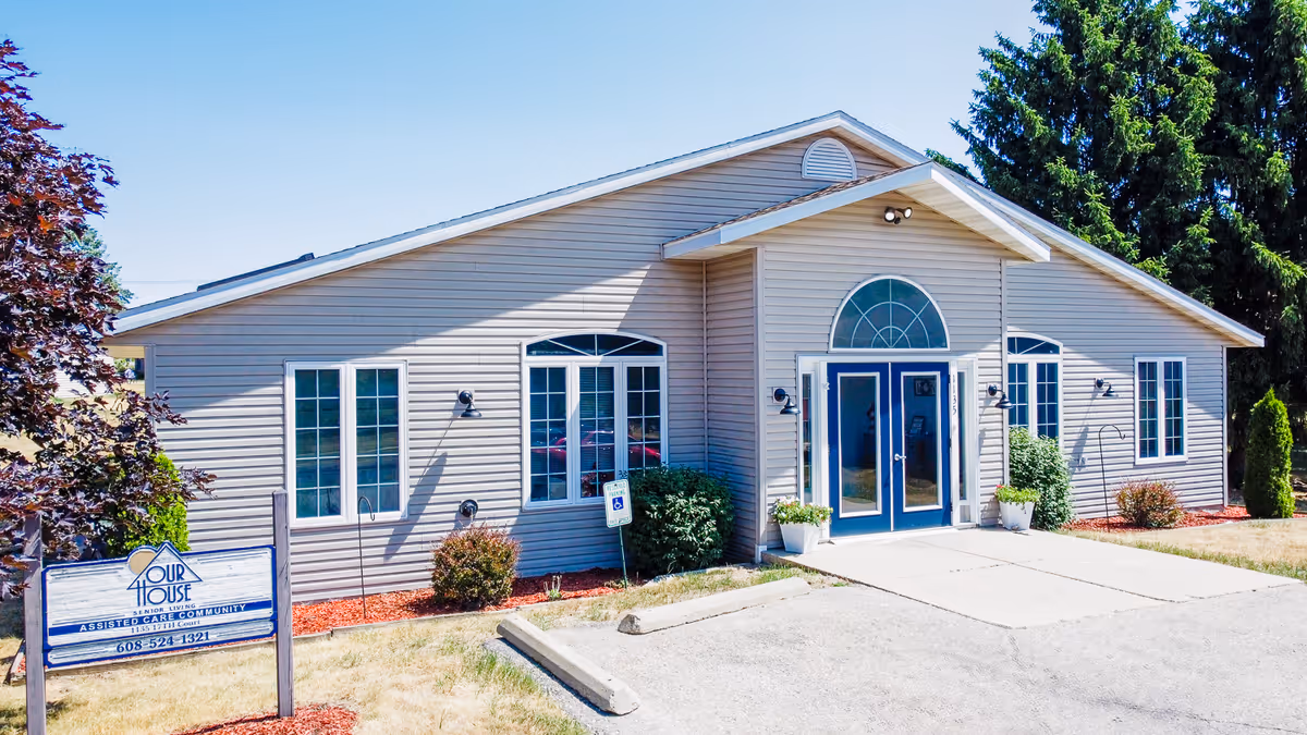 Exterior view of a single-story beige building with blue double doors and multiple windows, surrounded by small bushes and trees, with a sign in front that reads 'Our House Senior Living Assisted Care Community'.