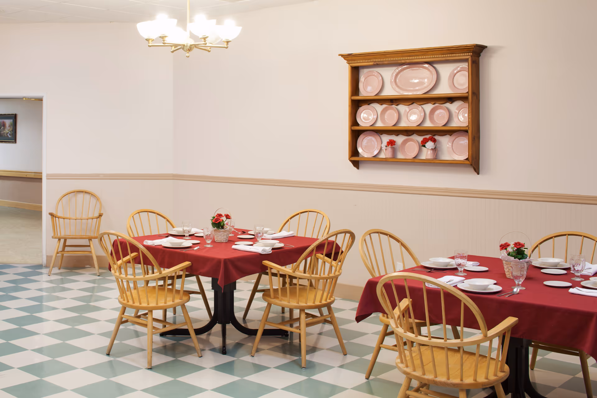 A dining room with two tables covered in red tablecloths, each set with white plates, glasses, and napkins. Wooden chairs surround the tables. A wooden wall shelf displays pink plates and a few small flower arrangements. The floor has a green and white checkered pattern, and a chandelier hangs from the ceiling.