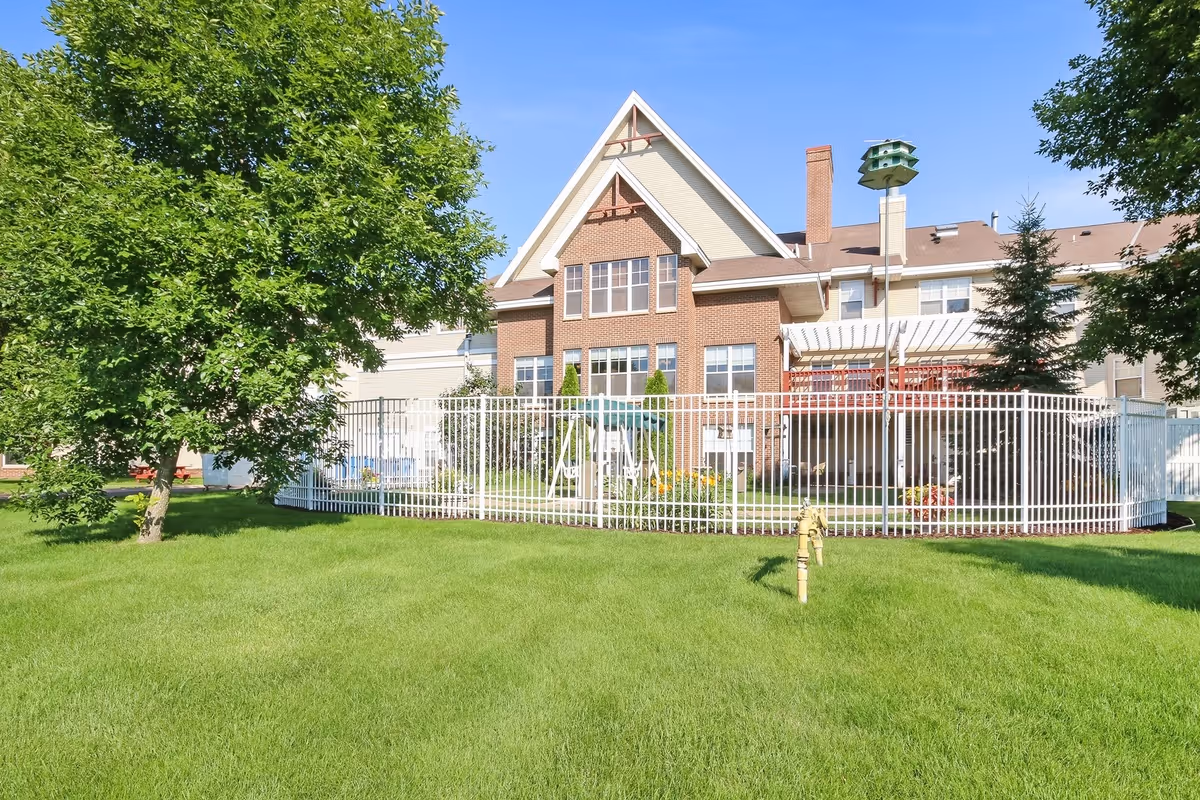 Front view of a brick-and-siding senior living building with a fenced grassy yard, trees, and a birdhouse.