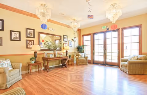 A bright and spacious living room area with wooden flooring, two striped armchairs with floral cushions, a wooden console table with decorative items and lamps, framed pictures on the wall, a large mirror, and glass double doors letting in natural light. The ceiling has three ornate chandeliers and some hanging decorations.