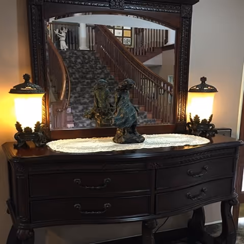 Dark wooden hallway console with mirror, two lit lamps, and a staircase reflected in the glass.