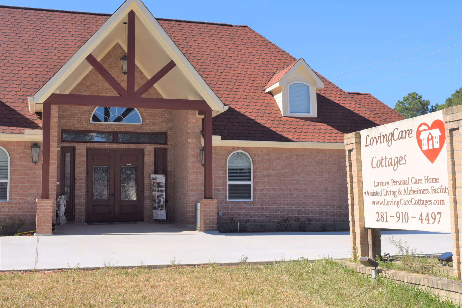 Exterior view of a brick building with a red roof and a covered entrance supported by wooden beams. There is a sign in front of the building that reads LovingCare Cottages, Luxury Personal Care Home, Assisted Living & Alzheimers Facility, with a website and phone number.