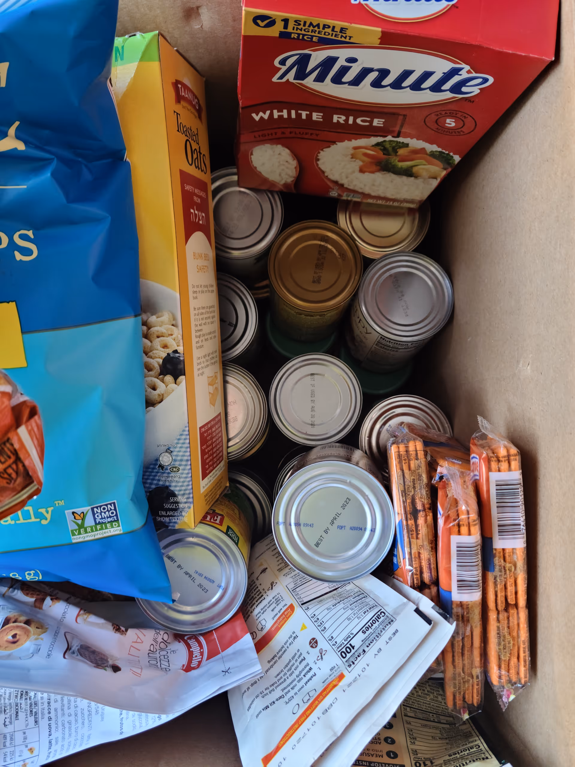 Boxed groceries including Minute Rice, oats, cans, and packaged snacks stacked inside a cardboard container.