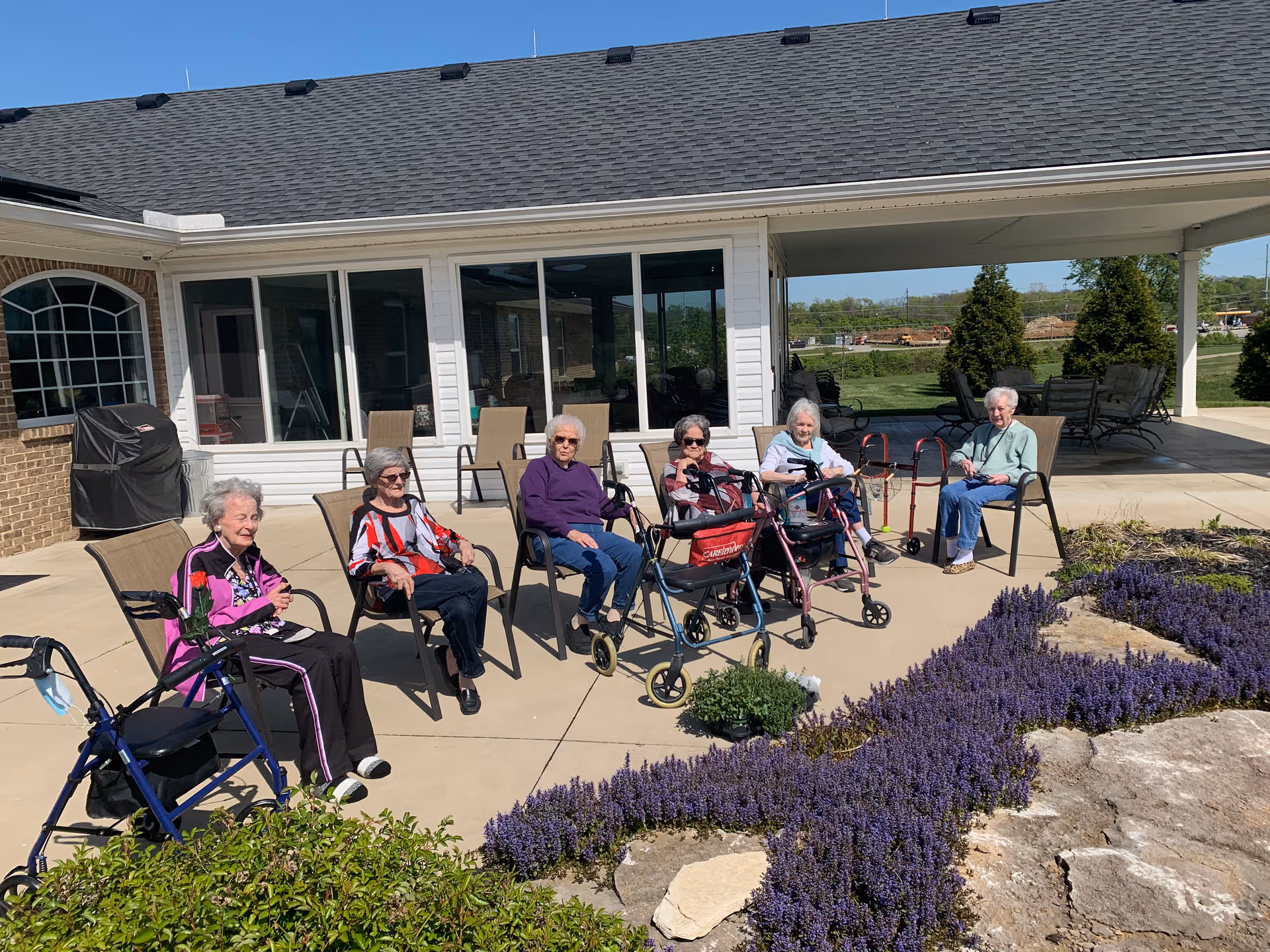 Six elderly women sitting outside on chairs on a sunny patio near a garden with purple flowers. Some women have walkers nearby. The building behind them has large windows and a covered area with additional seating.