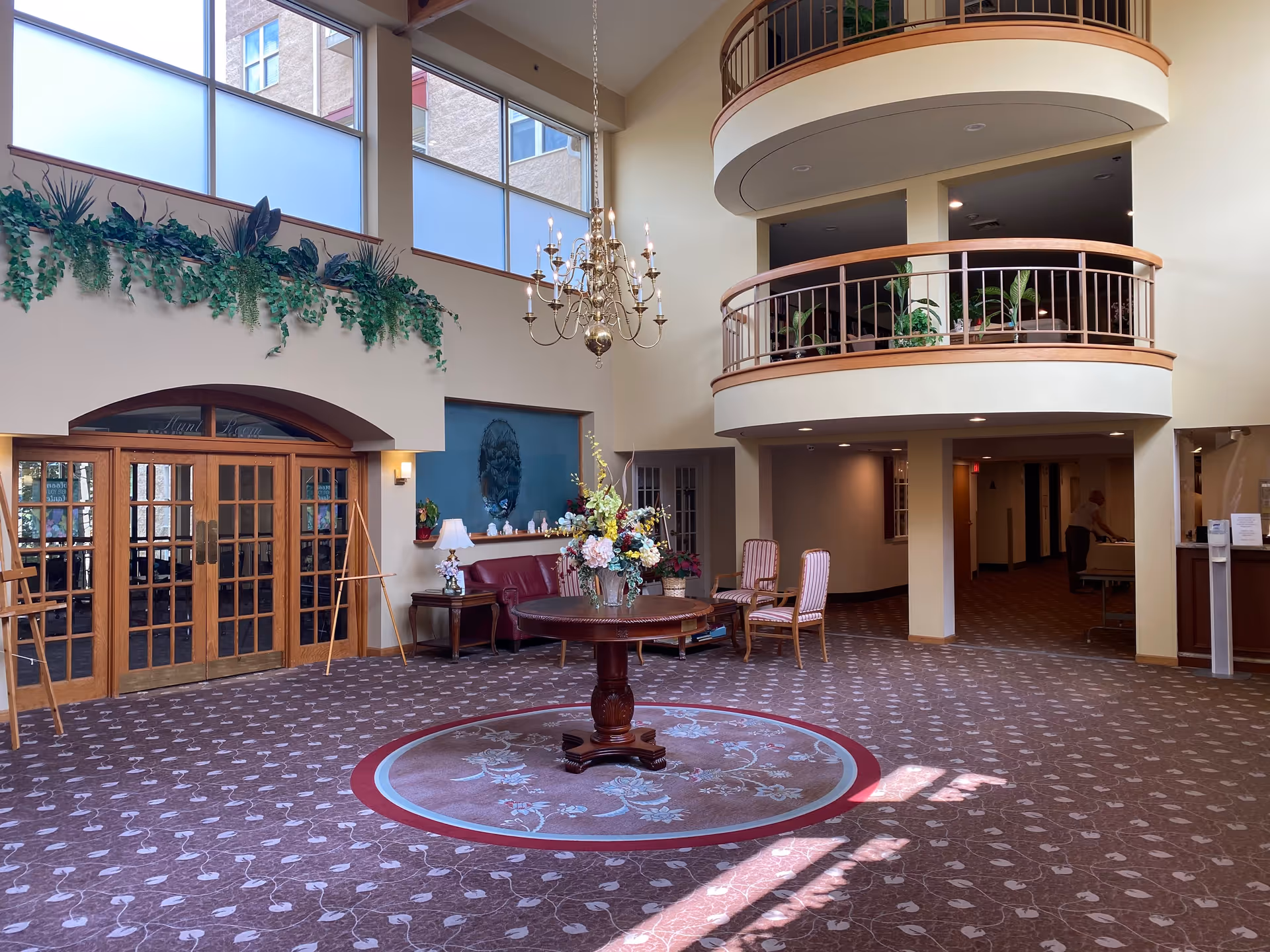 Spacious two-story lobby with a round central table topped with a floral arrangement, seating, balcony railings, chandelier, and large windows.