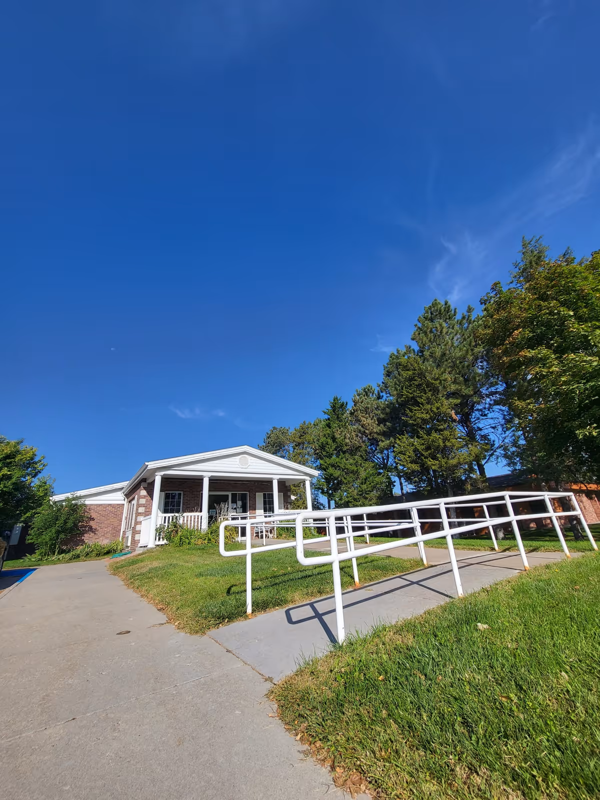 Exterior view of a single-story brick building with a white porch and railing, surrounded by green grass and trees under a clear blue sky. A concrete pathway with a white handrail ramp leads up to the entrance.