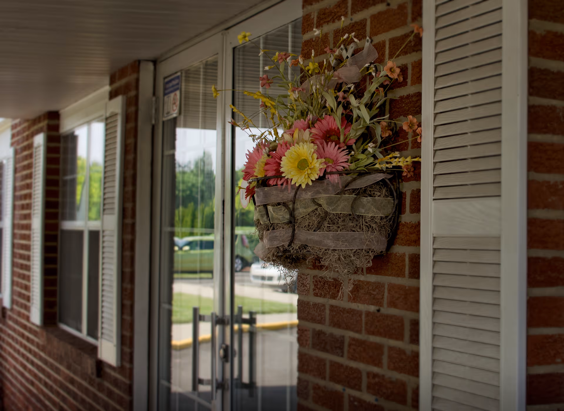 Close-up view of a brick wall with a decorative flower basket hanging on it, featuring pink and yellow flowers. To the left, there is a glass door with metal handles and windows with white shutters. The outdoor area and parked cars are visible through the glass door.