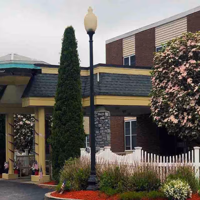 Covered entrance/porte-cochere of a senior living building with a lamppost, tall evergreen, white picket fence and flowering shrubs.