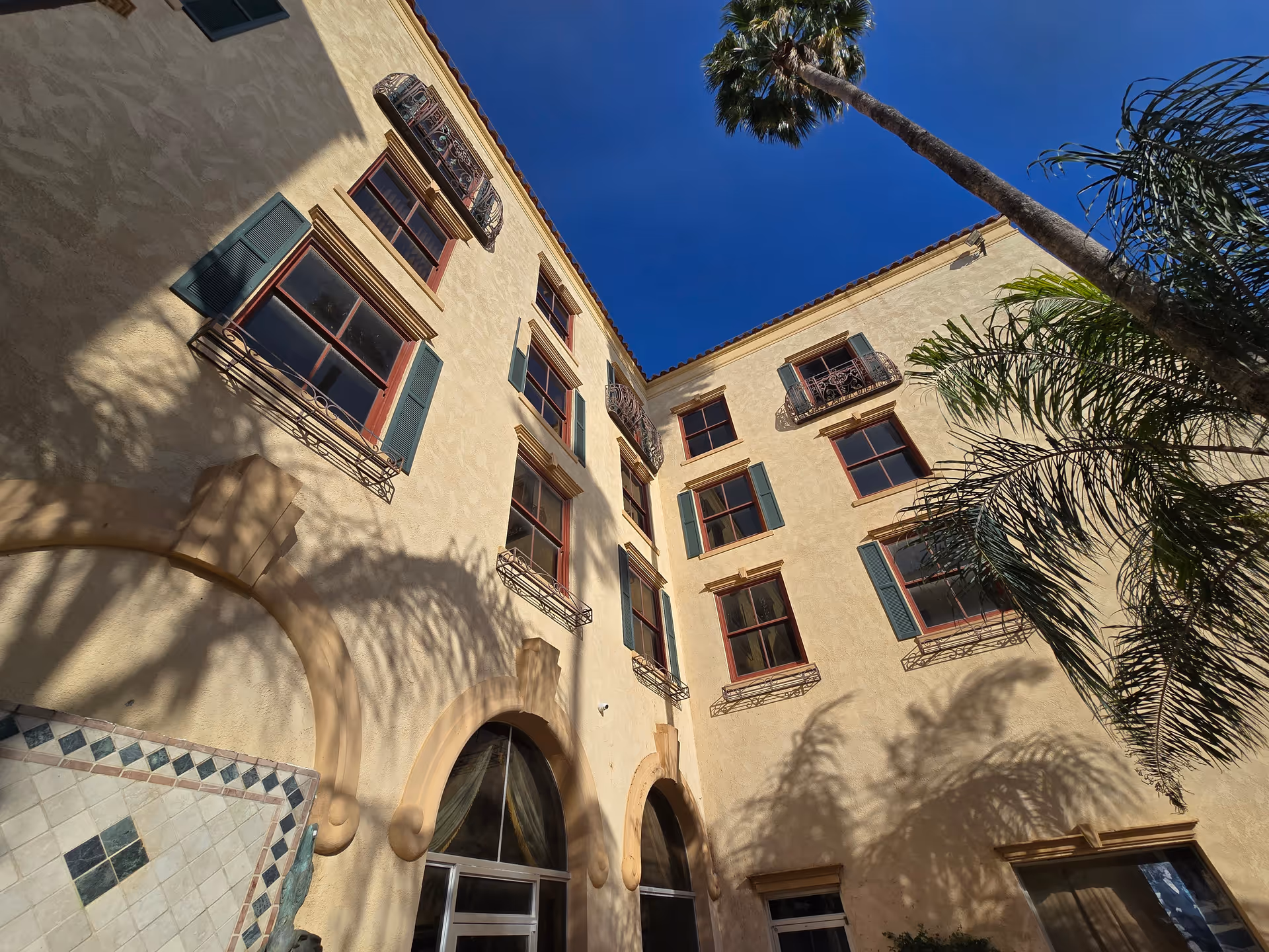 Exterior view of a tan stucco senior living building with arches, shutters, and palm trees.