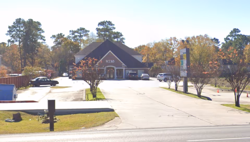 Exterior view of a senior care facility with driveway, parking, and autumn trees.