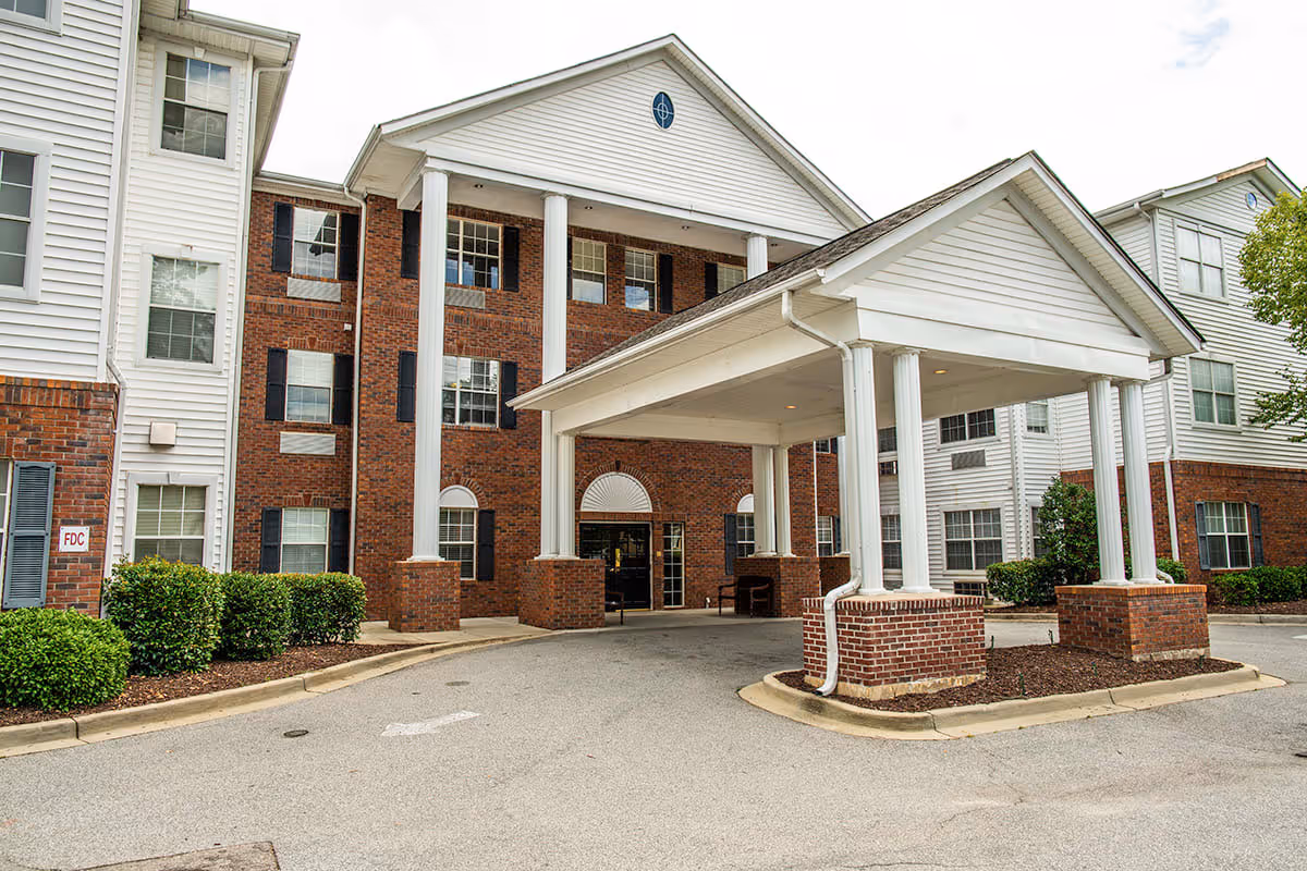 Front exterior view of a senior living facility building with a covered entrance supported by white columns and brick bases. The building has a combination of red brick and white siding with multiple windows and black shutters. There are neatly trimmed bushes along the driveway.