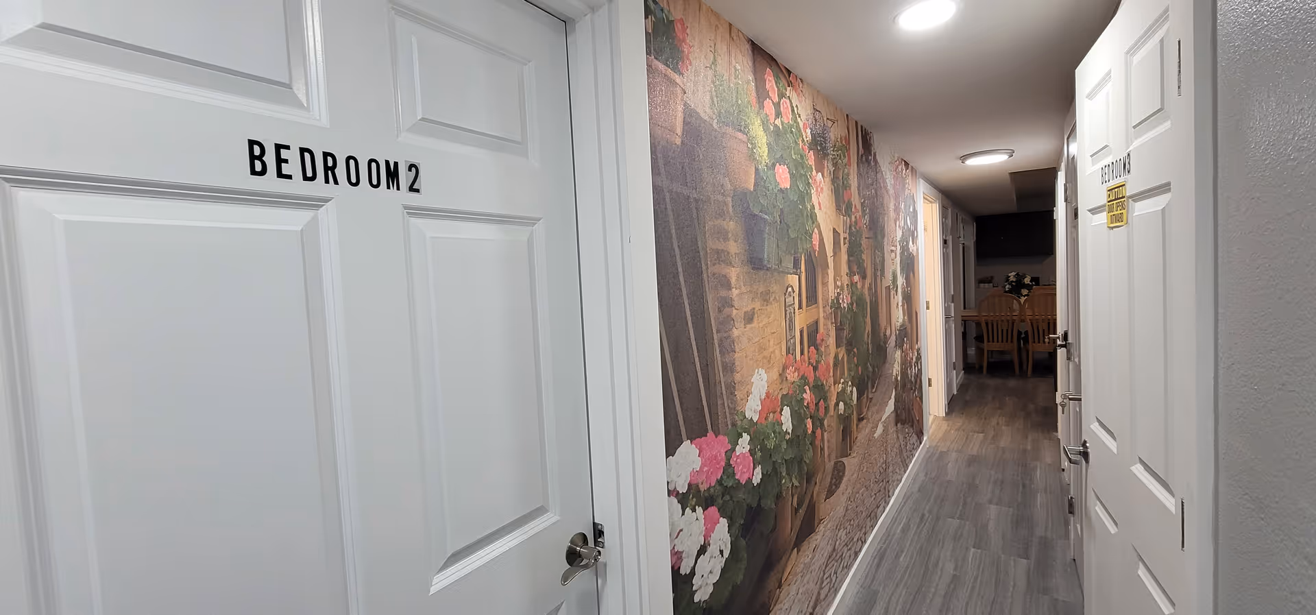 White doors line a floral hallway, leading to a dining area in a senior living facility.