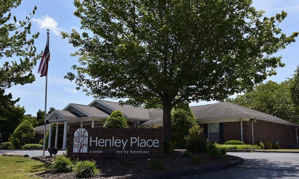 Front exterior of Henley Place senior living facility showing a brick building, entrance sign, large tree and an American flag.
