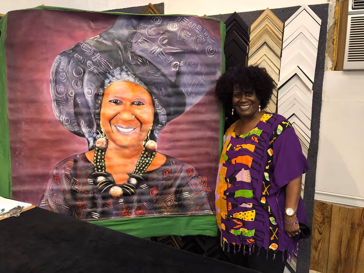 A smiling woman in a colorful dress stands beside a large portrait banner indoors.