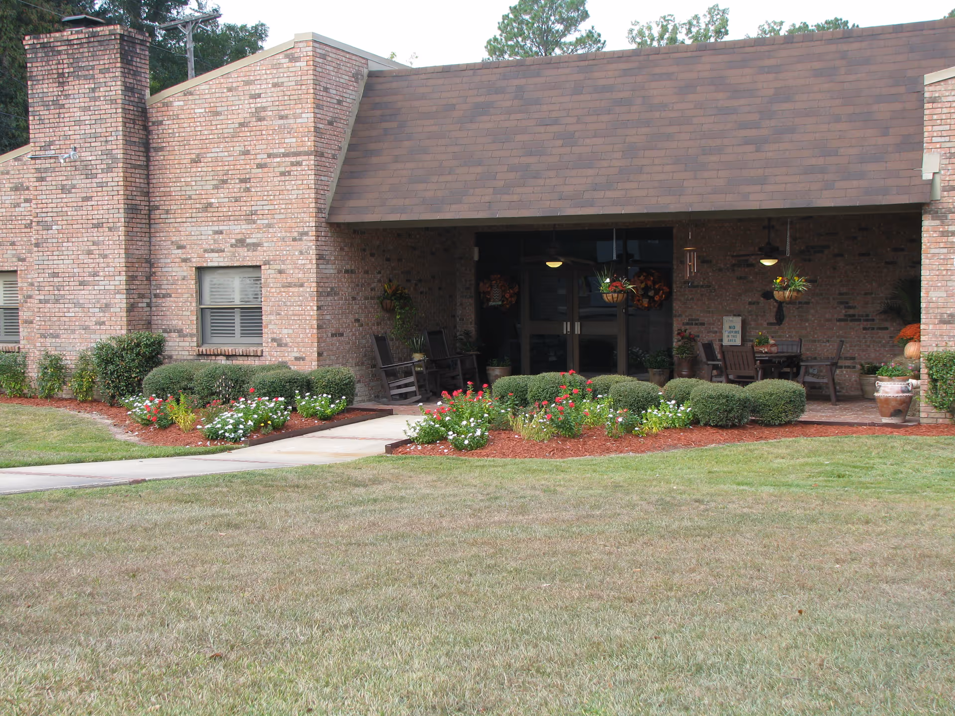 Exterior view of a brick building with a covered patio area featuring outdoor seating, hanging plants, and flower beds with shrubs and colorful flowers in front. A concrete walkway leads to the entrance.