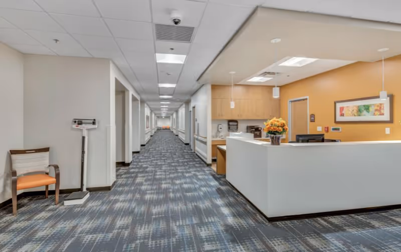 A long hallway in a healthcare facility with patterned carpet flooring, white walls, and ceiling lights. On the right side, there is a reception desk with a computer, a vase of orange flowers, and a framed picture on the wall behind it. On the left side, there is a chair and a medical scale. The hallway extends into the distance with doors and handrails along the walls.