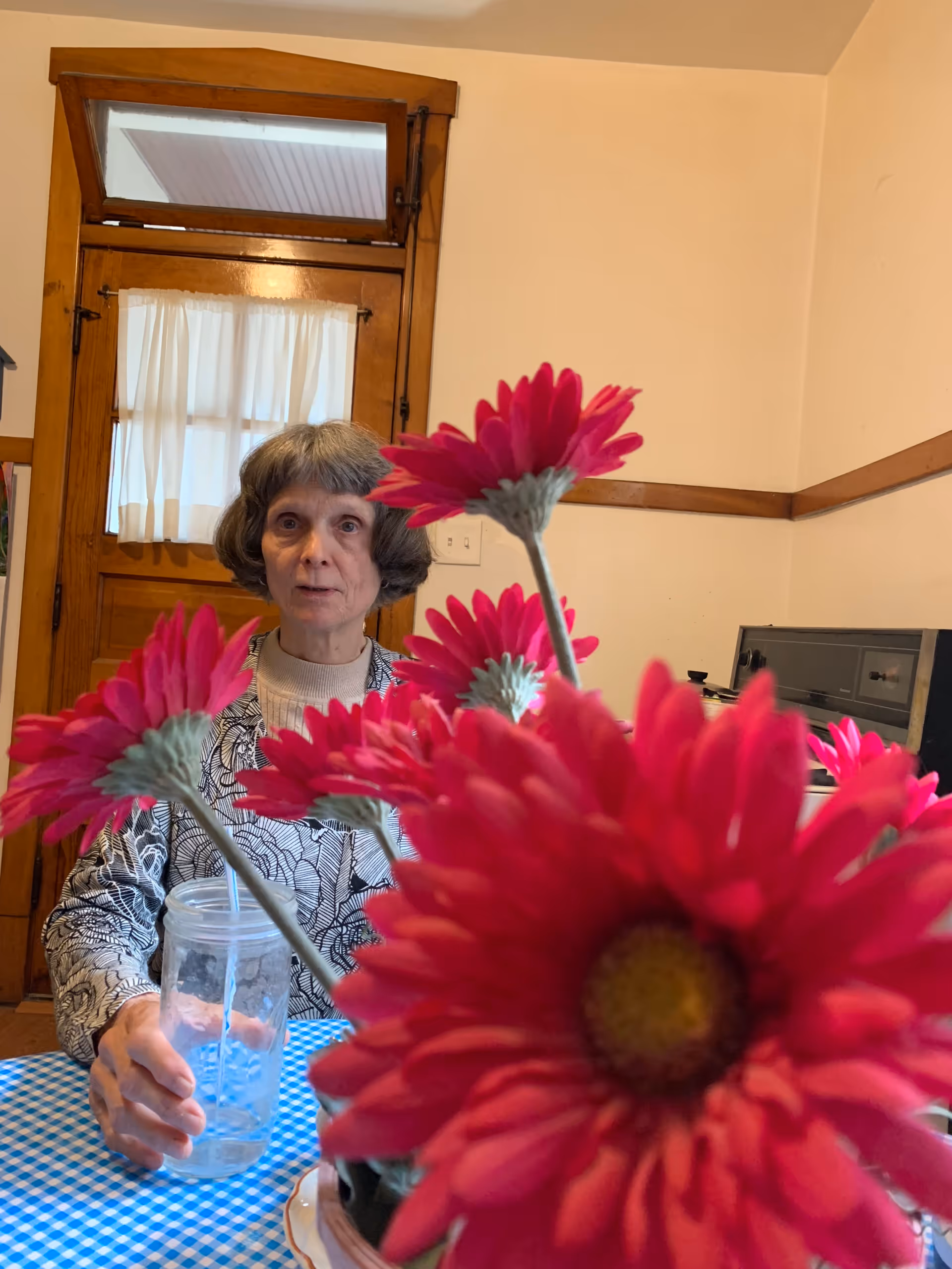 An elderly woman sits at a checkered table with pink flowers in foreground.
