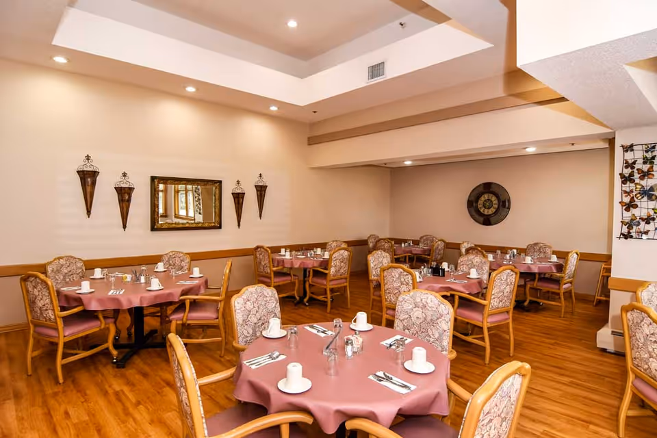 Dining room with round tables set with pink tablecloths and upholstered chairs under recessed lighting.