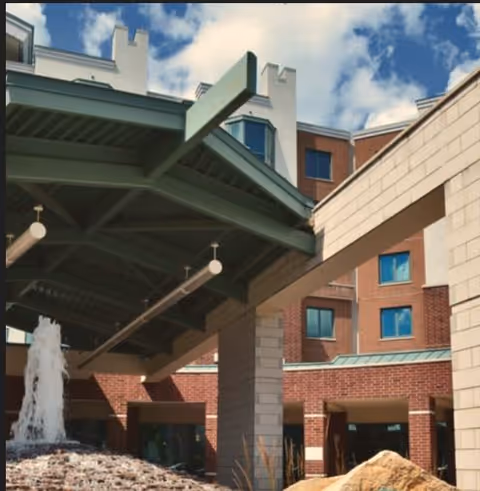 Covered brick entrance with a green metal canopy, a fountain and a multi-story building facade under a blue sky.